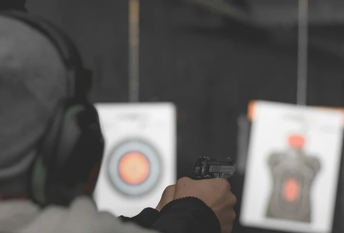 Person aiming for a target in an indoors shooting range. The focus is on the person, who is viewed from behind, with safety earmuffs, a black jacket and a gray beanie.