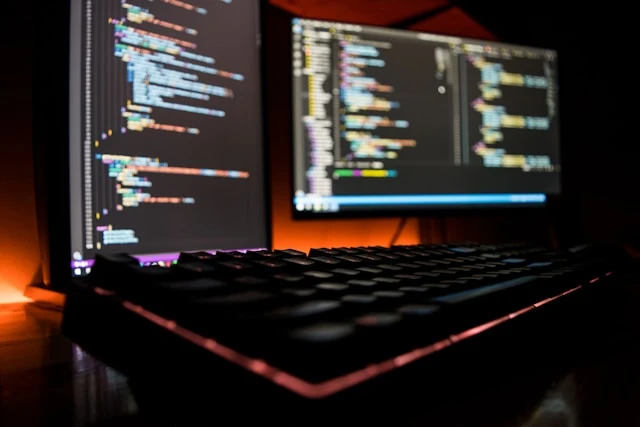A desk with two computer screens and a keyboard. The screens are blurry but display coding programs.